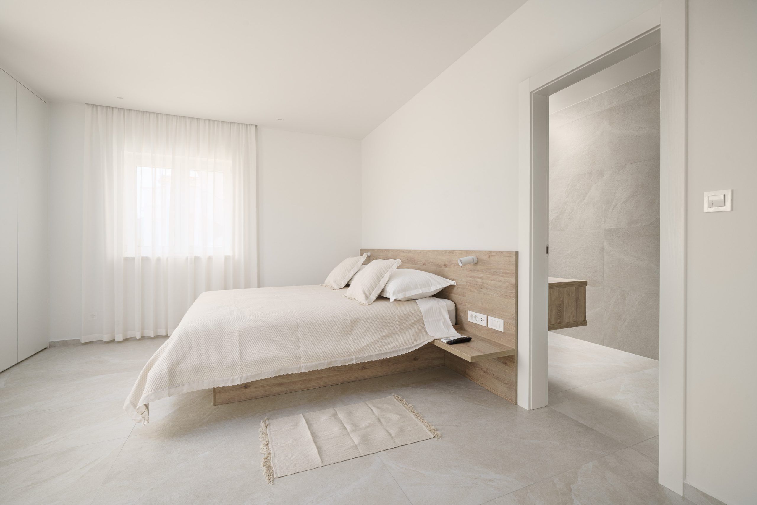Una camera da letto dal design minimalista, con un letto, un comodino e una finestra con tende leggere.