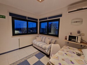 A room with a bed, a small table, a microwave, and two windows with a view of the outside. - Apartment Adri (Rovinj)