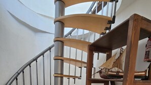En spiraltrappa med metallräcken och trästeg, som leder upp till en liten plattform med en trähylla där olika föremål står.