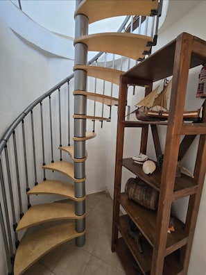 Eine Wendeltreppe mit Metallgeländern und Holzboden, die zu einer Zwischenebene führt, auf der sich ein Holzregal mit verschiedenen Gegenständen befindet.