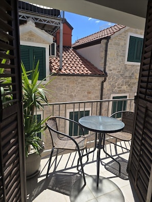 A balcony with a table and chairs, a potted plant, and a stone building with green shutters in the background. - House Vodice (Vodice)