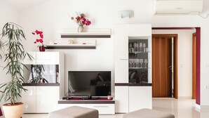 A modern living room with a flat-screen TV, a dining table, and a kitchen area with a refrigerator and shelves. - Apartments Villa Marija (Seget)