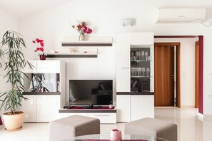 A modern living room with a flat-screen TV, a dining table, and a kitchen area with a refrigerator and shelves. - Apartments Villa Marija (Seget)