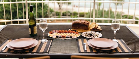 Una mesa de comedor dispuesta con platos, cubiertos, una botella de vino y una hogaza de pan, con vista a una masa de agua con barcos.