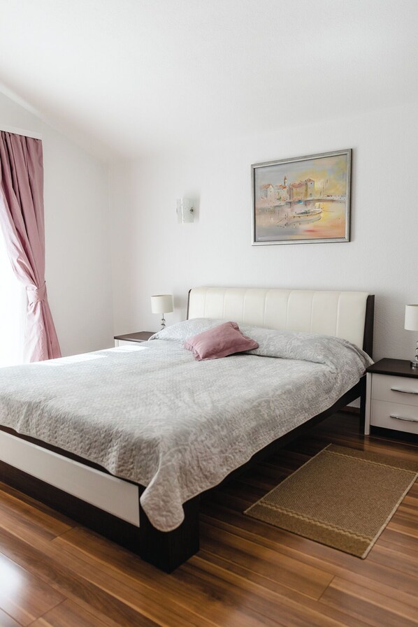 A bedroom with a bed, nightstands, a bedside lamp, a painting, a window with curtains, and a wooden floor. - Apartments Villa Marija (Seget)