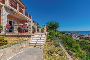 A stone building with a balcony, a staircase leading to the entrance, and a view of a coastal town. - Apartments Andrija (Rab)