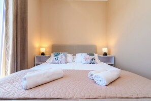 A neatly made bed with a quilted headboard, two bedside tables with lamps, and a window with curtains. - Apartments Andrija (Rab)