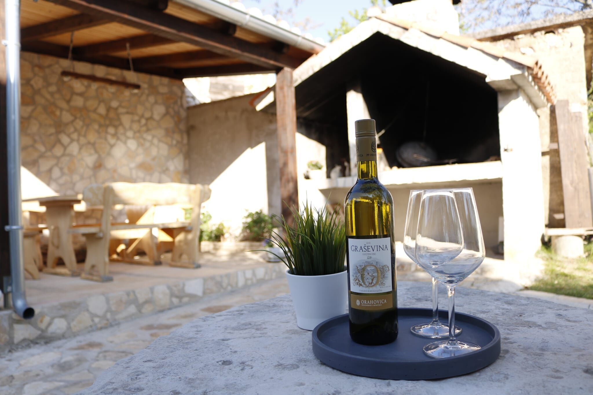 A bottle of wine and a glass on a tray, with a stone wall and outdoor seating area in the background.