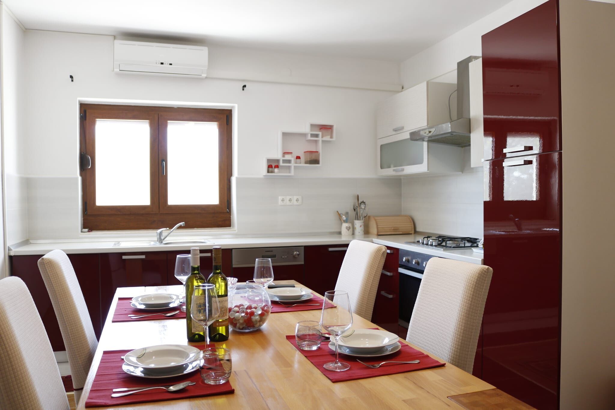 Un coin repas avec une table en bois dressée pour quatre personnes, une fenêtre offrant une belle vue et une cuisine dotée de meubles rouges et d’appareils électroménagers blancs.