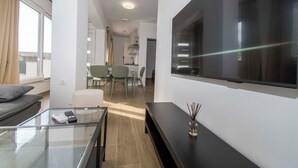 A modern living room with a glass coffee table, a sofa, a flat-screen TV mounted on the wall, and a dining area in the background. - Apartments Stay Near Paklenica (Starigrad)