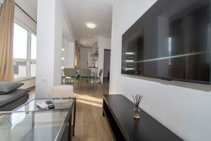 A modern living room with a glass coffee table, a sofa, a flat-screen TV mounted on the wall, and a dining area in the background. - Apartments Stay Near Paklenica (Starigrad)