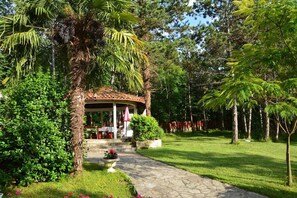 A covered outdoor seating area with a stone pathway leading to it, surrounded by lush greenery and trees. - Apartment Villa Zizi (Viskovo)