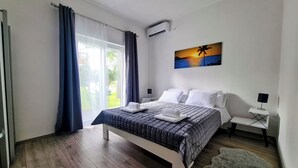 A bedroom with a bed, a painting of a sunset, a window with curtains, and an air conditioning unit. - House Beachfront Delight (Pasman)