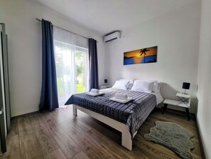 A bedroom with a bed, a painting of a sunset, a window with curtains, and an air conditioning unit. - House Beachfront Delight (Pasman)