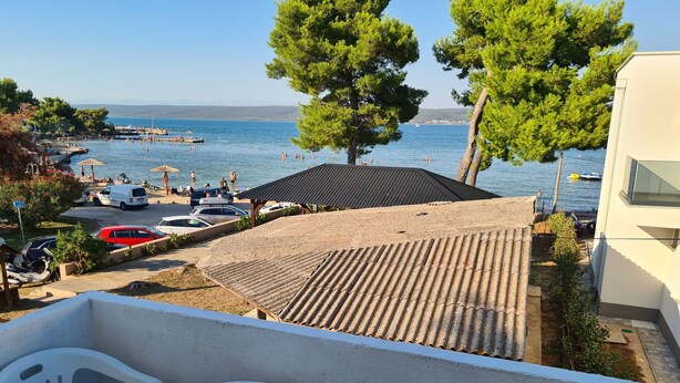 A balcony with two white plastic chairs and a small round table, overlooking a beach with people swimming and boats in the water. - House Beachfront Delight (Pasman)