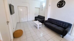 A living room with a white door, a television, a black sofa, a white coffee table, and a round orange ottoman. - House Beachfront Delight (Pasman)