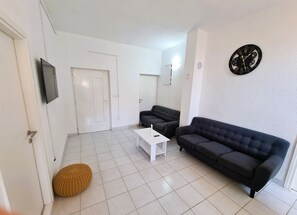 A living room with a white door, a television, a black sofa, a white coffee table, and a round orange ottoman. - House Beachfront Delight (Pasman)