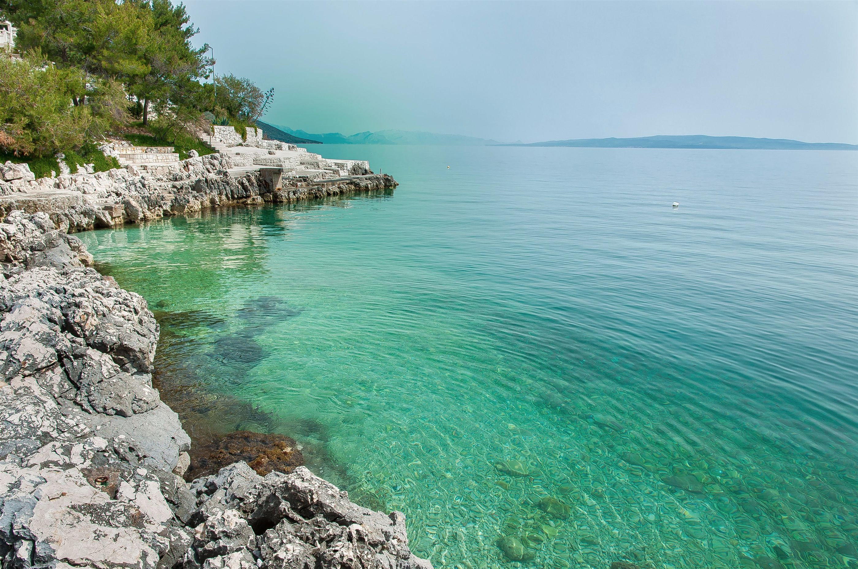 A coastal landscape with clear turquoise waters, rocky shoreline, and distant mountains.