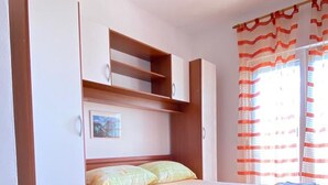A bedroom with a wooden bed, built-in storage, a window with curtains, and a wooden floor. - Apartments Mia (Crikvenica)