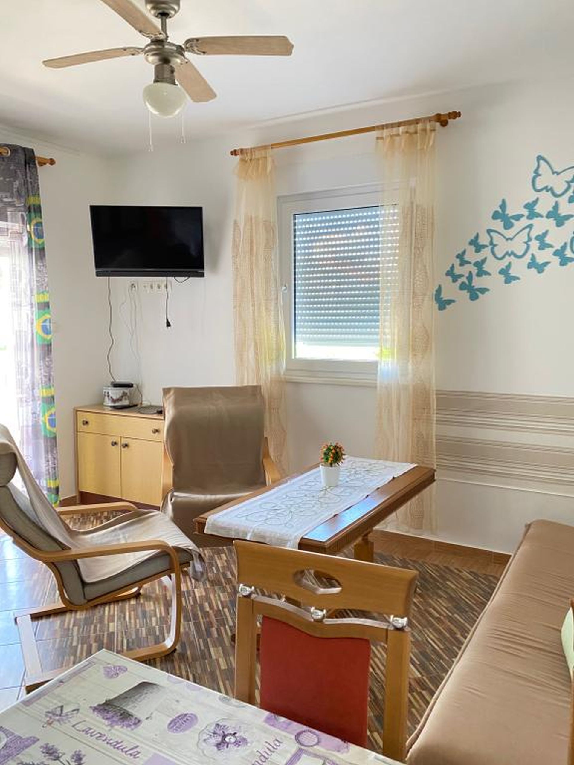 A room with a ceiling fan, a television, a window with blinds, a table with a tablecloth, and a wall with butterfly decals.