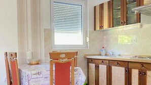 A dining area with a table and chairs, a window with blinds, and a kitchen with wooden cabinets. - Apartments Mia (Crikvenica)