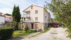 A multi-story building with a gravel driveway, a parked car, and a garden area with shrubs and a tall tree. - Apartment Ljubica (Rab)