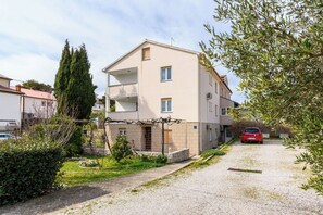A multi-story building with a gravel driveway, a parked car, and a garden area with shrubs and a tall tree. - Apartment Ljubica (Rab)