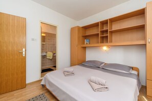 A bedroom with a bed, a wooden wardrobe, a shelf unit, and a bathroom visible through an open door. - Apartment Ljubica (Rab)