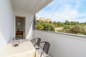 A balcony with a table and chairs, a door leading to an interior room, and a view of buildings and greenery. - Apartment Ljubica (Rab)