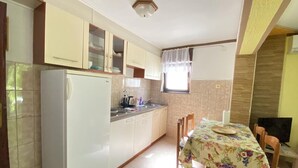 A compact kitchen with a refrigerator, cabinets, a dining table with chairs, and a window with curtains. - Apartments Fran (Crikvenica)