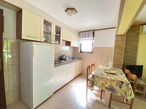 Eine kleine Küche mit Kühlschrank, Schränken, einem Esstisch mit Stühlen und einem Fenster mit Vorhängen.