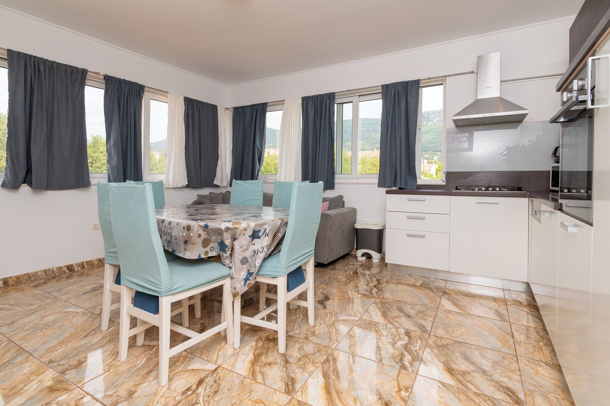 A modern kitchen with a dining area, marble flooring, and large windows with curtains.