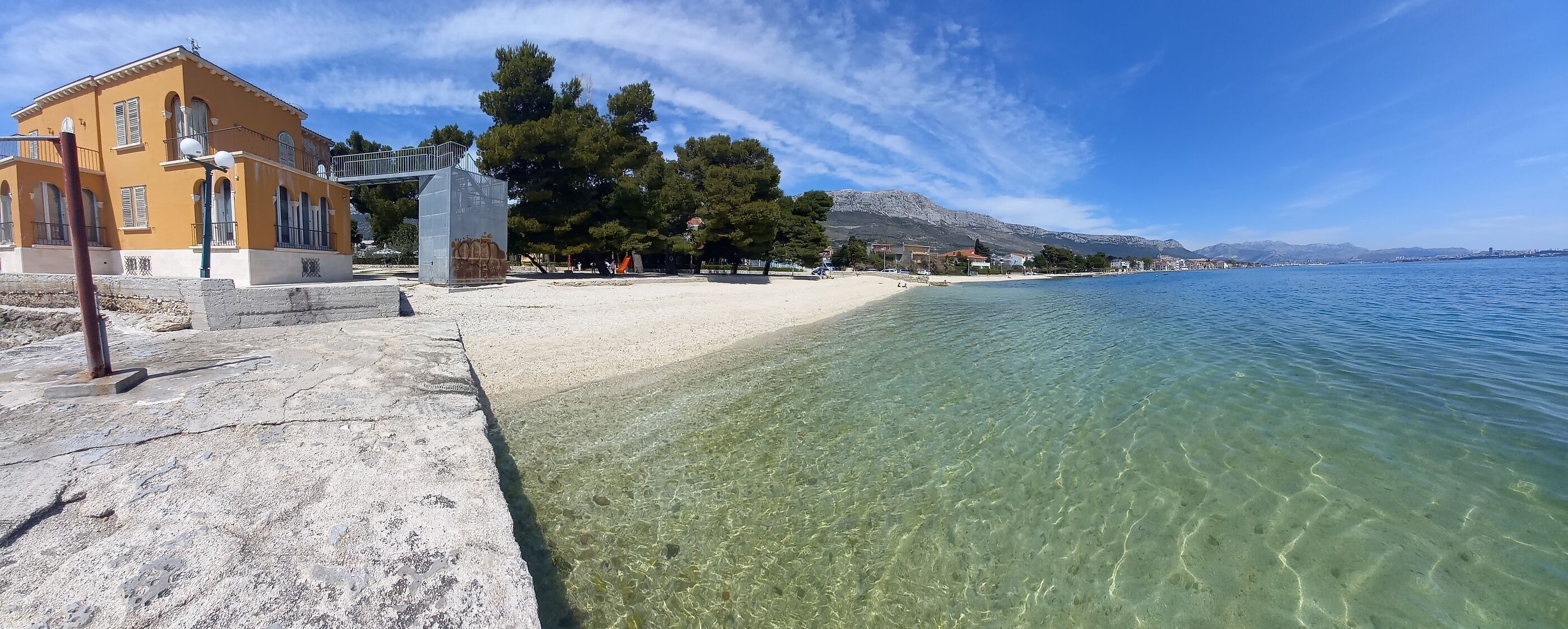 A coastal scene with a clear beach, a yellow building, and a mountain in the background.