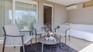 Een moderne hotelkamer met een bed, twee fauteuils, een tafeltje met een schaal fruit en een fles, en een groot raam met gordijnen.