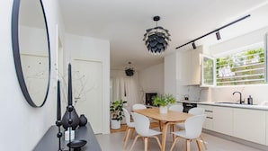 Een moderne keuken met witte kasten, een houten eettafel en witte stoelen. Er hangt een grote spiegel aan de muur, een hanglamp en een raam met uitzicht op groen.