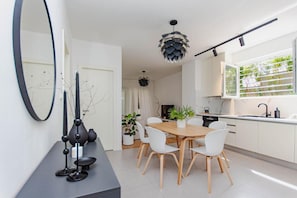 A modern kitchen with white cabinets, a wooden dining table, and white chairs. There is a large mirror on the wall, a hanging pendant light, and a window with a view of greenery. - Apartment Sea View Garden (Split)