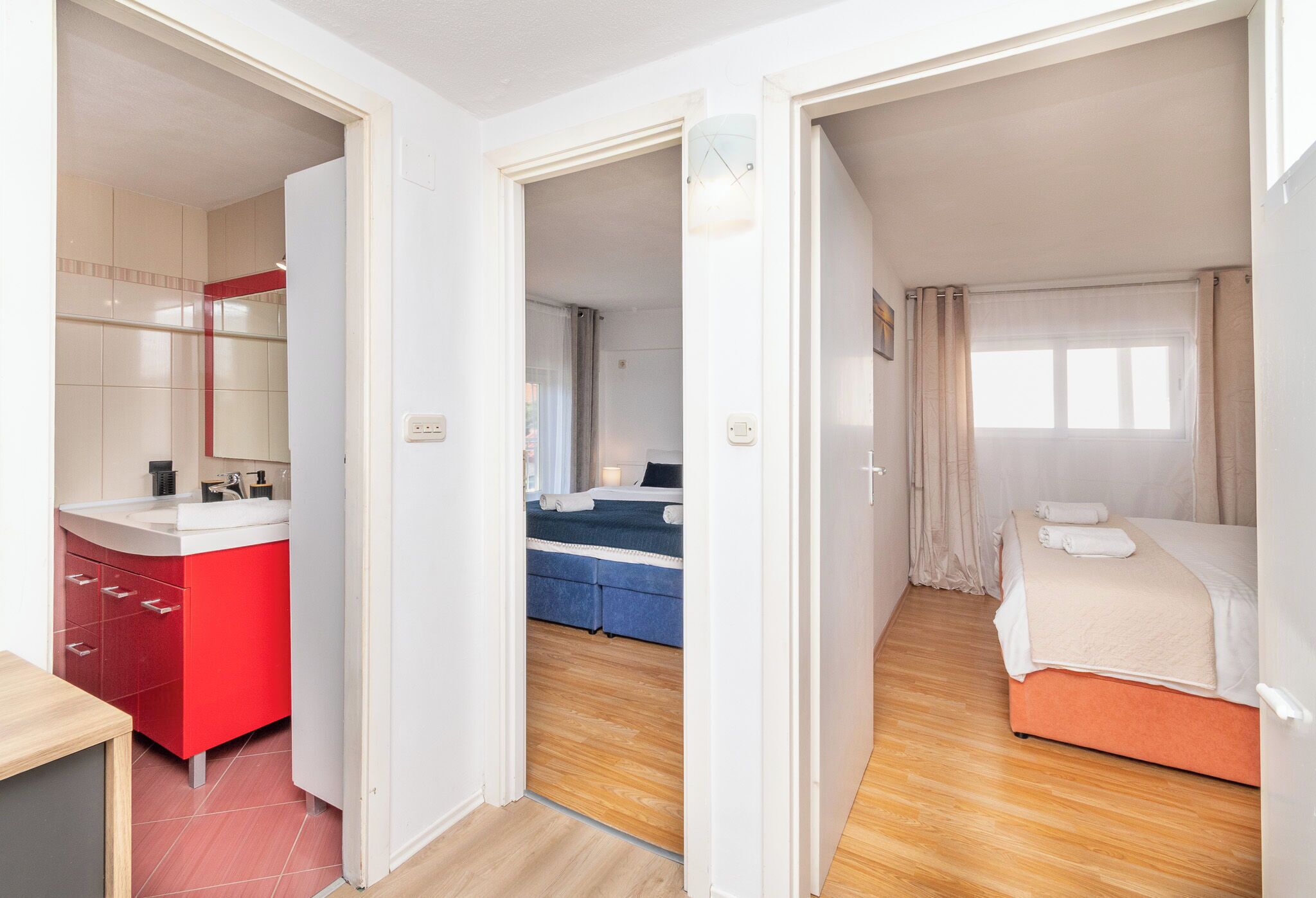 Ein Hotelzimmer mit Holzboden, einem Bett mit beiger Decke, einem Badezimmer in Rot und Weiß sowie einem Blick nach draußen durch ein Fenster.