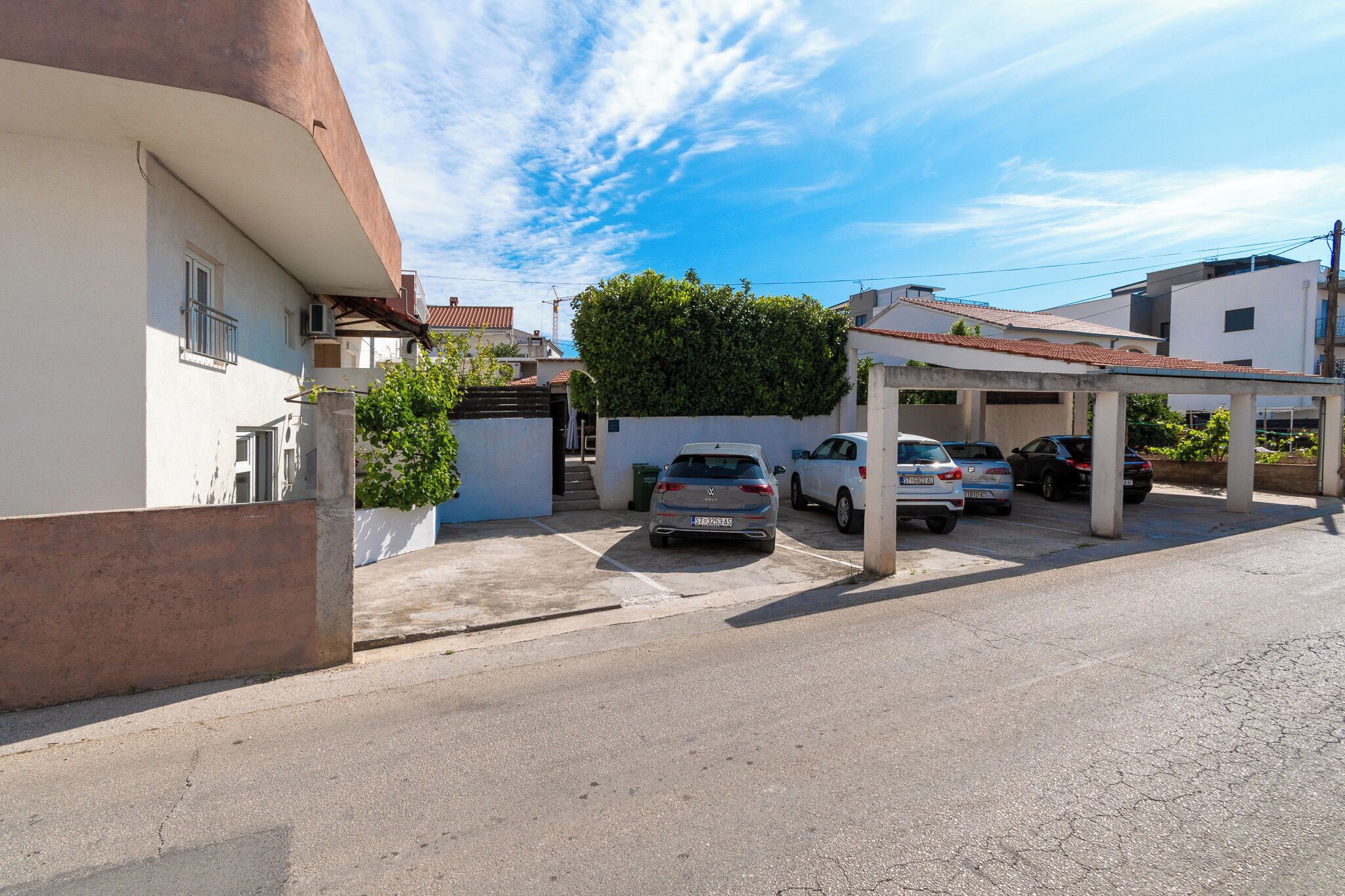 A residential street with parked cars, a covered parking area, and houses with visible balconies and windows.