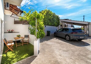 A car parked in a driveway, a small patio with a table and chairs, and a green hedge. - Apartment Dijana (Okrug)