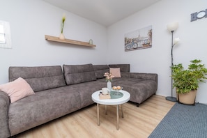 Ein modernes Wohnzimmer mit einer grauen Couch, einem kleinen runden Tisch mit einer Vase und einem Buch, einer Topfpflanze und einer Wandregal mit Dekorationen.