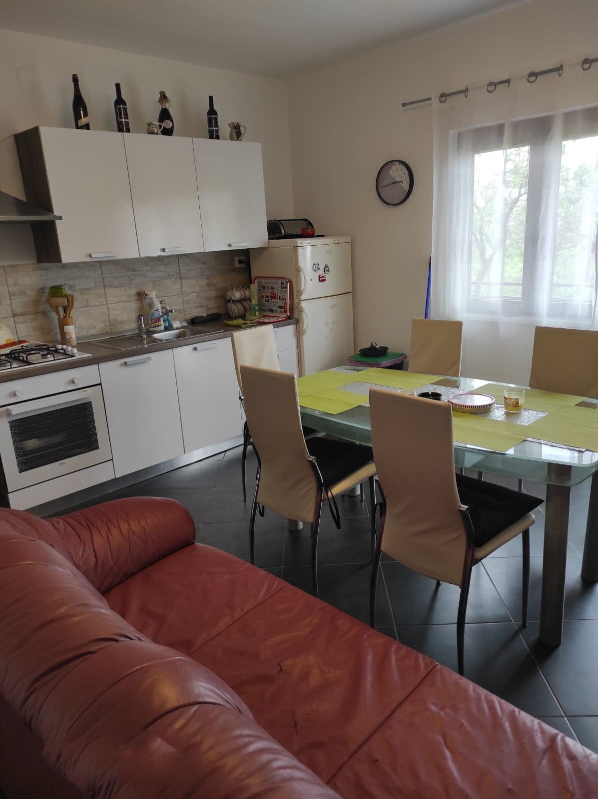 Eine moderne Küche mit weißen Schränken, einem Esstisch mit Stühlen, einem roten Ledersofa und einem Fenster mit Vorhängen.