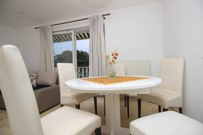 Ein kleiner, gut beleuchteter Raum mit einem runden Tisch, vier weißen Stühlen, einem Sofa und einem Fenster mit Vorhängen.