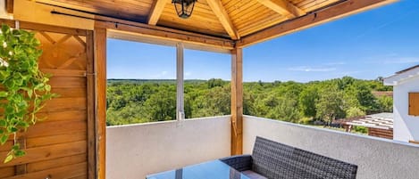 Un balcón exterior con una mesa de vidrio y sillas de mimbre, techo de madera y vistas a un paisaje boscoso.