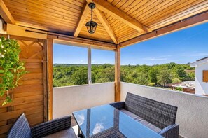 Ein Balkon mit Gartenmöbeln aus Rattan, einem Glastisch und Blick auf eine bewaldete Landschaft. Die Decke ist aus Holz.
