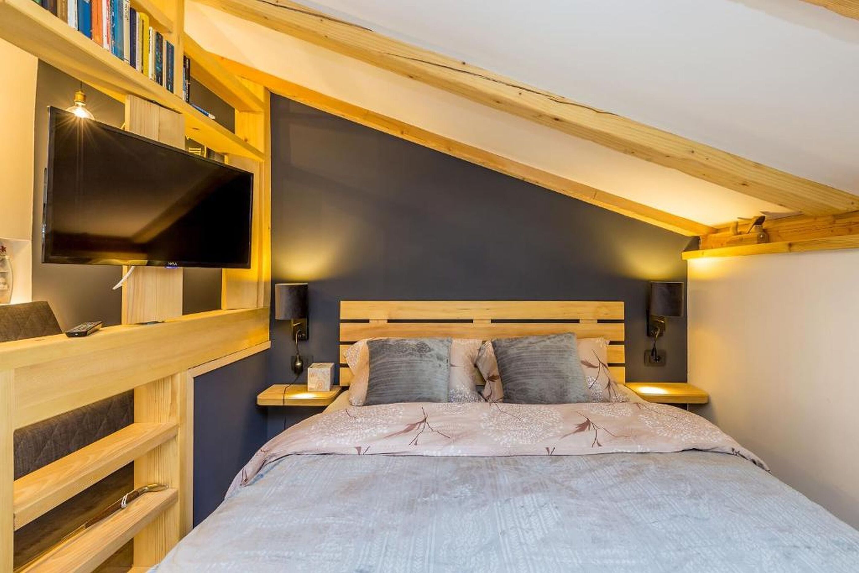 A bedroom with a bed, wooden headboard, bedside tables, a TV, and bookshelves.