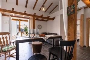 Ein gemütliches Zimmer mit Holzbalkendecke, einem Esstisch mit Stühlen, einer Couch und Blick ins Freie.
