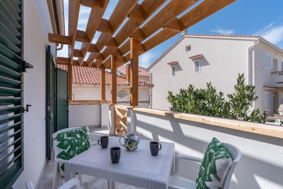 Un balcón con una pérgola de madera, una mesa blanca con sillas y vistas a edificios cercanos.
