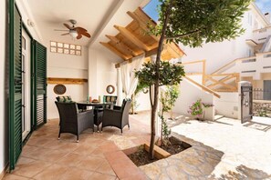 Eine überdachte Terrasse mit Tisch, Stühlen und einem Deckenventilator, sowie ein Baum.