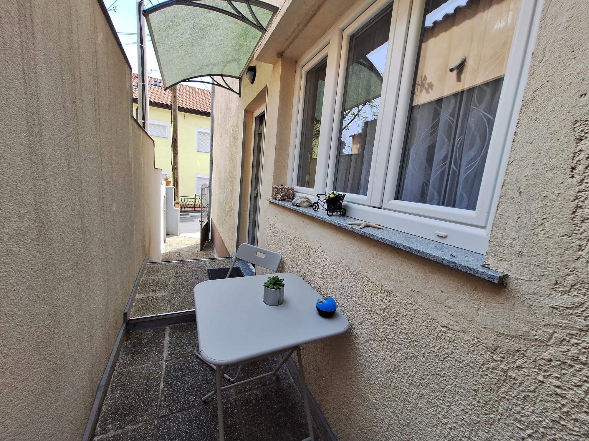 A narrow outdoor corridor with a white table and chairs, a small potted plant, and a window with a shelf above.