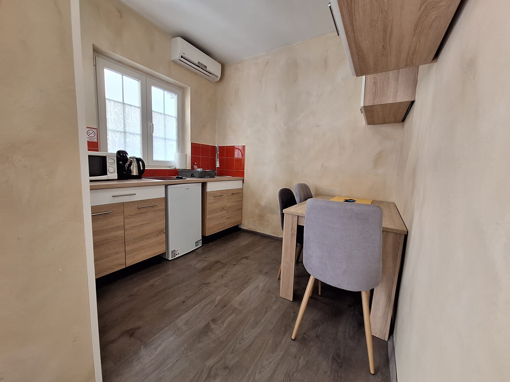 A compact kitchen with a microwave, coffee maker, and a small refrigerator. There is a dining table with two chairs and a window with blinds.